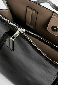 Interior de un bolso de cuero negro con un zipper plateado, forro beige y cierre a presión. Cuenta con una textura suave y detalles de costura mínimos.