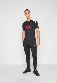 T-shirt de sport Nike noire avec le logo "AXA" rouge et le logo de Liverpool, associé à un jogging Nike noir et des chaussures de sport noires.