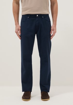 Homme portant un pantalon droit bleu foncé, une chemise beige rentrée, et des chaussures en daim marron, debout sur un fond clair.