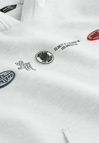 Sweat-shirt blanc en tissu texturé, avec divers écussons : un emblème de basketball, des coordonnées géographiques, et du texte stylisé.