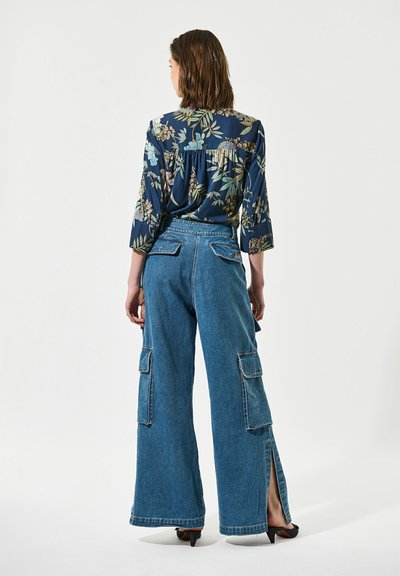 Femme debout, dos tourné, portant une blouse florale sombre à manches trois-quarts et un jean cargo bleu taille haute à jambes larges avec des fentes latérales.