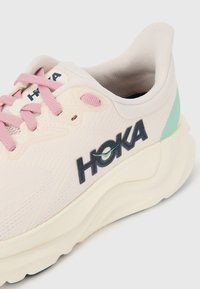 Light beige athletic shoe with pink laces and logo, textured fabric upper, white cushioned sole, and green accent on heel.