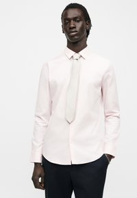 Άνδρας με σκουρόχρωμο δέρμα και πλεξούδες φορώντας ανοιχτό ροζ πουκάμισο, ανοιχτό γκρι γραβάτα και σκούρο παντελόνι, στέκεται μπροστά από ένα μονόχρωμο φόντο.