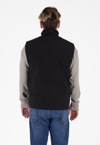 Gilet nero senza maniche sopra una maglietta a maniche lunghe grigia chiara, caratterizzato da un colletto alto e una vestibilità ampia, abbinato a jeans in denim blu.