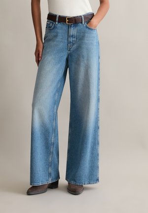 Wijde blauwe denim jeans met een lichte wassing, voorzien van voorzakken en een bruine riem met een rechthoekige gesp. Getextureerde stof.