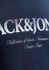 Navyblå T-shirt med hvidt trykt tekst: "JACK & JONES" og "Definition of denim. Nineteen m. Design Dept." i forskellige skrifttyper.