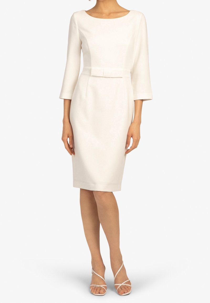 Femme portant une robe blanche ajustée jusqu'aux genoux avec des manches trois-quarts et un petit nœud à la taille, assortie à des talons blancs à brides.