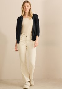 Veste zippée navy sur un haut beige, assortie à un pantalon large crème et des baskets blanches, montrant un look décontracté et moderne.