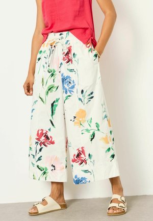 Woman wearing white wide-leg pants with colorful floral print, a red sleeveless top, and beige buckle sandals, standing against a white wall.