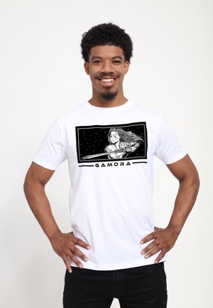 Biała koszulka z bawełny z czarnym nadrukiem graficznym Gamory z mieczem na tle gwiaździstego nieba. Na dole znajduje się napis "GAMORA".