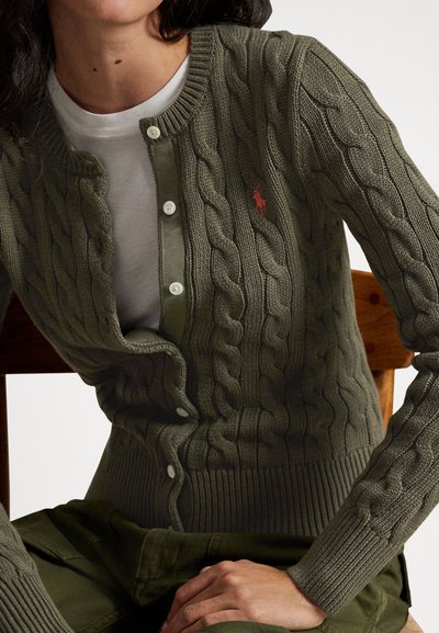 Καρντιγκάν από ελαιί πράσινο πλεκτό ύφασμα με στρογγυλή λαιμόκοψη, διακοσμημένο με λευκά κουμπιά και ένα μικρό κόκκινο λογότυπο. Ριμπ στις άκρες και τις μανσέτες με υφαντό σχέδιο.