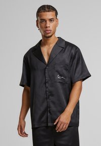 Schwarzes Satin-Hemd mit kurzen Ärmeln, ausgestattet mit einem Schlitzkragen, einer Brusttasche und einem gestickten Akzent. Glatte Textur mit subtilen Mustern.