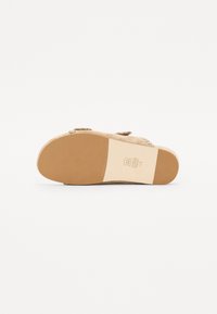 Sandale slide en suede beige avec une semelle lisse et un dessus texturé comportant deux lanières. Marquage et étiquette visibles sur la semelle.