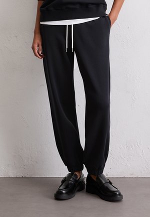 Pantalons de jogging noirs avec des poignets élastiques, dotés d'une taille à cordon crème. Le matériau semble doux avec une texture lisse.