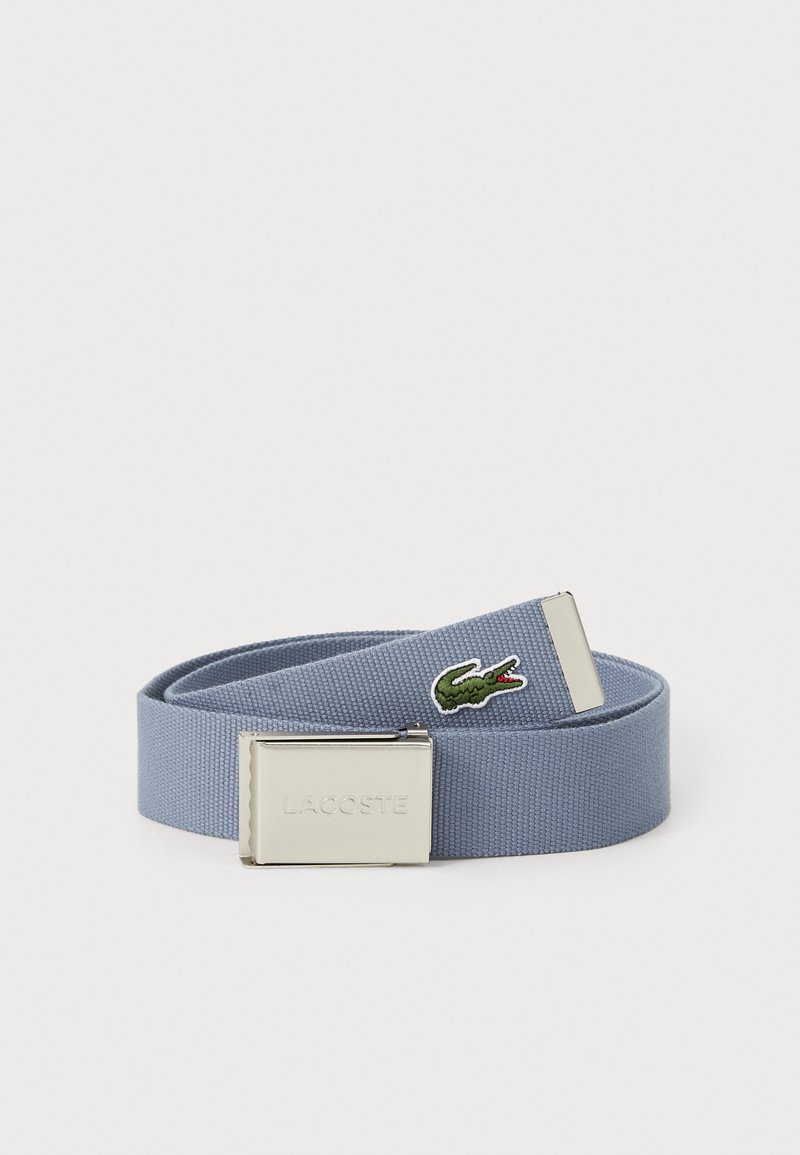 Blå vevet stoffbelte med en sølvfarget metallspenne, med en brodert grønn krokodille-logo og merkevarenavnet "LACOSTE."