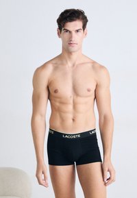 Homem homem sem camisa com cabelo curto e ondulado, vestindo boxers pretos da Lacoste, de pé contra um fundo claro e liso.