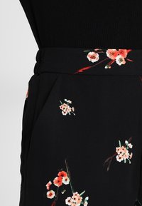 Tissu noir à motif floral avec des fleurs pêche et blanches. Texture lisse avec une ceinture. Vue rapprochée d'un pantalon.