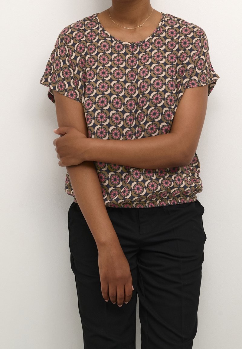 Personne portant un chemisier à manches courtes avec des motifs floraux circulaires en rose, beige et noir, assorti à un pantalon noir, debout contre un mur blanc.