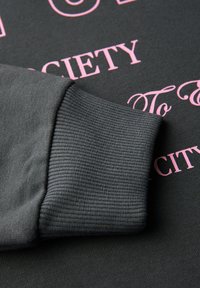 Felpa grigia con polsini a costine e texture morbida, con testo rosa stampato su uno sfondo scuro.