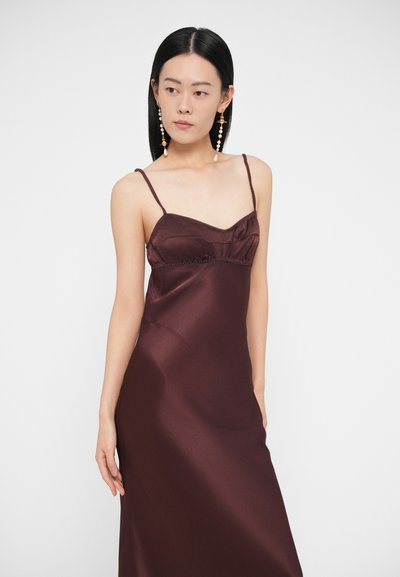 Žena s rovnými čiernymi vlasmi má na sebe tmavofialové saténové šaty na tenkých ramienkach a dlhé ozdobné perlové visiace náušnice, pozerá sa doľava.