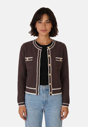 Mujer con un cárdigan marrón oscuro texturizado con ribete crema y botones dorados, sobre una camiseta negra y vaqueros azul claro, de pie frente a un fondo blanco.