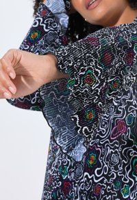 Schwarzes, plissiertes Kleid mit bunten Blumen- und abstrakten Mustern. Mit Rüschensäumen und transparenten Details sowie kontrastierenden Stoffstrukturen.