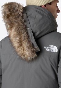 Grå vattentät jacka med en fuskpälsluva, med en vit logotyp på vänster axel. Närbild från baksidan och sidan.