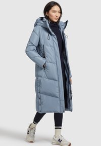 Cappotto lungo imbottito di colore blu chiaro con cappuccio, zip frontale, due tasche laterali e dettagli con bottoni a pressione lungo le cuciture laterali. Indossato con scarpe sportive.