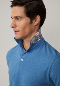 Modrá polokošile s texturovaným povrchem, s vzorovaným límcem v zeleném a burgundském květinovém designu. Jednoduché zapínání na knoflík.