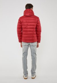 Giacca a vento rossa con cappuccio, progettata con sezioni trapuntate. Il modello indossa jeans grigi chiari e scarpe casual, vista da dietro.