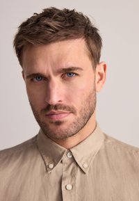 Beiges Hemd zum Knöpfen aus weichem Stoff, mit Kragen und feiner Textur. Das Modell hat kurze, zerzauste dunkle Haare und Bartstoppeln.