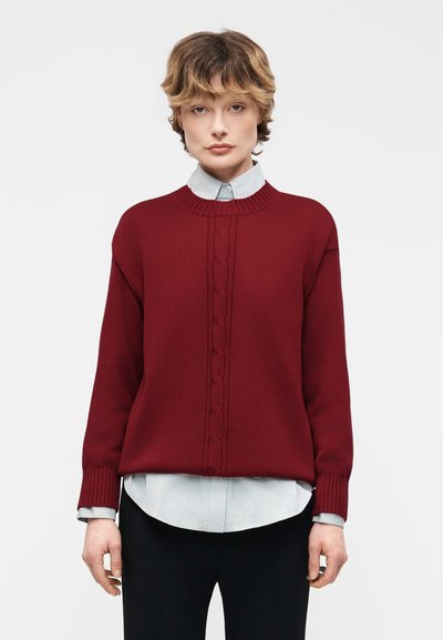 Jeune personne aux cheveux courts portant un pull rouge en maille torsadée par-dessus une chemise rayée à boutons et un pantalon noir, debout sur un fond blanc.