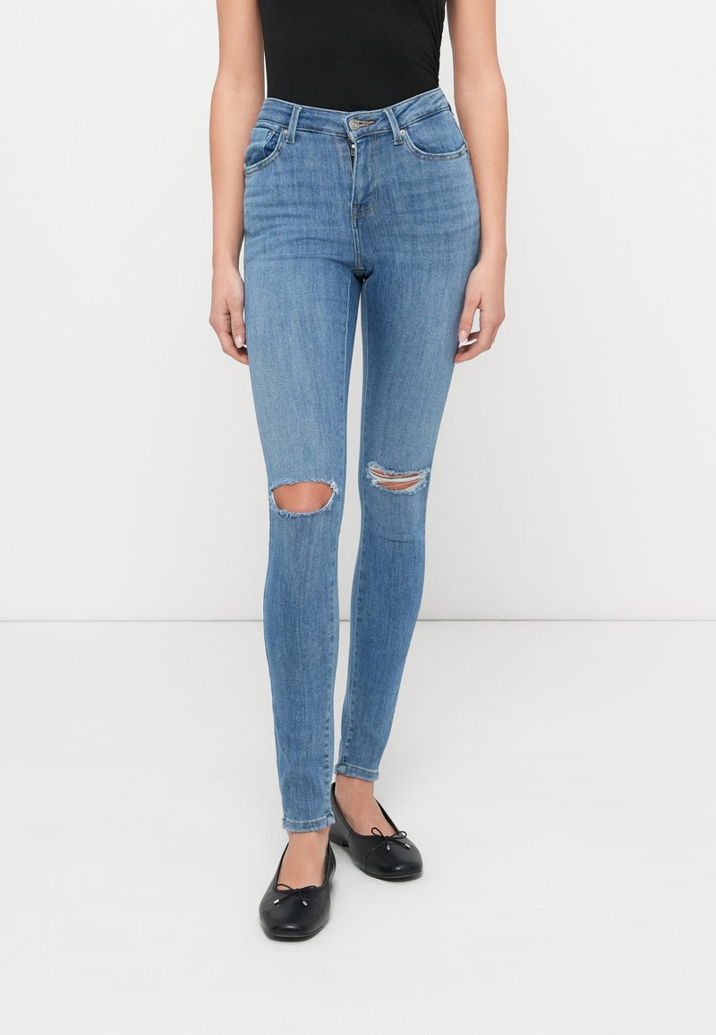 Λεπτά μπλε τζιν από ελαστικό denim με ψηλή μέση, σκισίματα στα γόνατα και κλασικό σχέδιο με πέντε τσέπες. Συνδυάζονται με μαύρα επίπεδα παπούτσια.