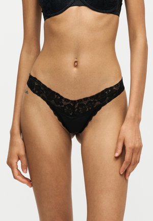 Femme portant un soutien-gorge en dentelle noire et un string assorti en dentelle noire, debout devant un fond uni et clair.