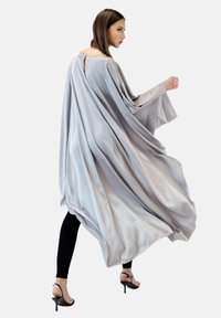 Fließender, hellgrauer Umhang aus glattem Stoff, mit weiten Ärmeln, einem Rückenschlüssellochdetail, kombiniert mit schwarzen Leggings und Sandalen.