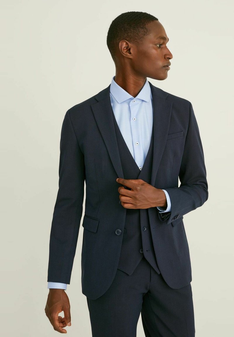 Marinblå kostym med ett strukturerat mönster, enfärgad jacka och matchande väst. Ljusblå skjorta under, som visar en slät textur.