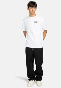 Hvid bomulds t-shirt med sort grafisk tekst på venstre bryst, parret med sorte denim bukser og hvide sneakers, stående mod en ensfarvet baggrund.
