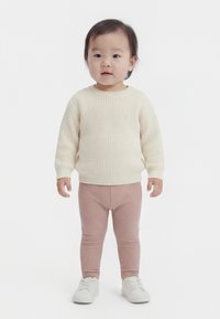 Børnehavebarn stående iført en cremefarvet strikket sweater, lyserøde ribstrikkede leggings og hvide sneakers mod en ensfarvet hvid baggrund.