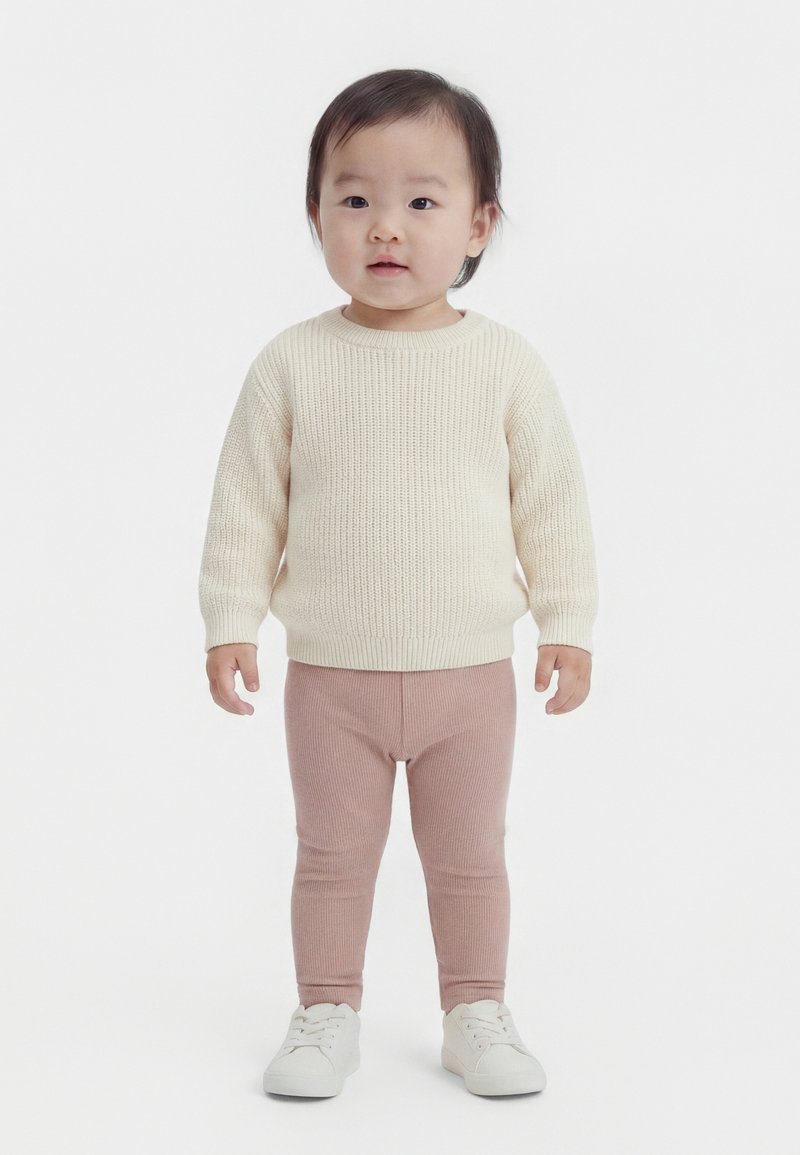 Børnehavebarn stående iført en cremefarvet strikket sweater, lyserøde ribstrikkede leggings og hvide sneakers mod en ensfarvet hvid baggrund.