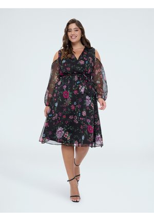Lachende vrouw die een zwarte midi-jurk met bloemenprint draagt met doorschijnende cold-shoulder mouwen en zwarte enkelriem hakken loopt naar voren.