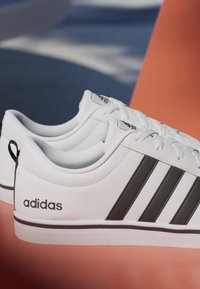 Zapatillas Adidas blancas con rayas negras y logo, colocadas junto a un cono de tráfico naranja sobre una superficie azul.