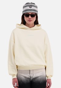 Vrouw in een crèmekleurige hoodie, zwart-witte gradient jeans, grijs gestreepte beanie met logo, zwarte zonnebril en zilveren ringoorbellen.