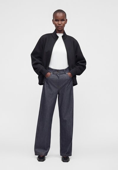 Personne portant une veste noire, un haut blanc, un pantalon large gris foncé et des chaussures noires, debout les mains dans les poches.