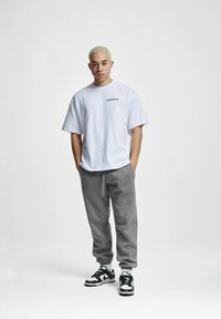 Biała bawełniana koszulka z czarnym logo, w zestawieniu z szarymi dresami. Model nosi czarno-białe sneakersy. Prosty design z luźnym krojem.