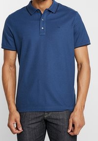Polo-shirt bleu marine en coton, avec un col classique, une patte de boutonnage à trois boutons et un logo sur le côté gauche de la poitrine. Manches courtes.