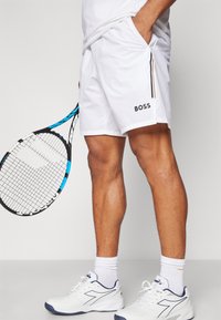 Pantaloni sportivi bianchi con logo nero "BOSS", tasche laterali e strisce arancioni e nere. Il giocatore tiene una racchetta da tennis blu e nera.