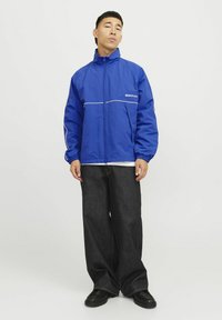 Blå vindjakke med lynlås, hvide detaljer og en afslappet pasform. Sammen med løse mørke jeans og kraftige sorte sko.