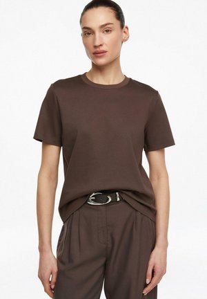 Kobieta w ciemnobrązowej koszulce z krótkim rękawem włożonej do pasujących plisowanych spodni, nosząca czarny pasek ze srebrną klamrą, stojąca na białym tle.