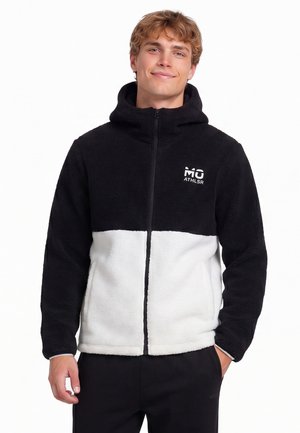Junger Mann trägt eine schwarz-weiße Fleecejacke mit Reißverschluss und Kapuze, steht mit einer Hand in der Tasche und lächelt leicht.
