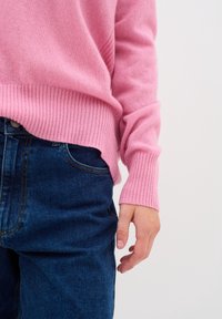 Różowy sweter z kaszmiru z prążkowanymi mankietami i dołem, zestawiony z ciemnoniebieskimi dżinsami. Sweter ma luźny krój i miękką fakturę.